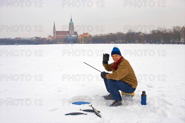 A man sits ice fishing on the Theresienwiese, with three freshly caught mackerel, waving to the