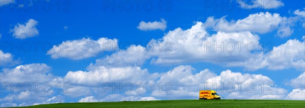 A DHL delivery van on a country road in the middle of meadows with blue sky and many white fair