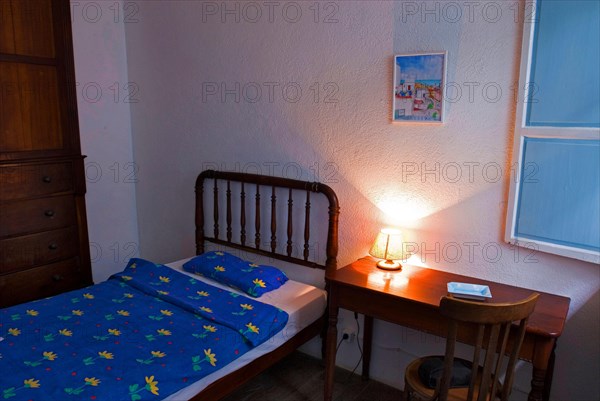 Bed and wooden table with illuminated bedside lamp in the room of a small, simple hotel or