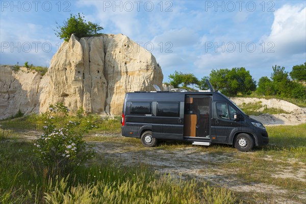 A motorhome stands in front of a rocky formation under a blue sky with clouds, mountain with