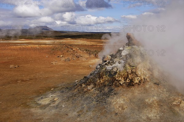 Close-up of steaming vent or fumarole in a geothermal landscape with a view of distant hills at