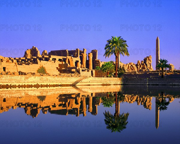 Palm trees grow beside the cistern, obelisk, columns and other remains at temple of Karnak, Luxor,