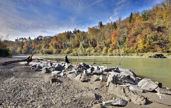 An angler sits on large stones by a river in autumn, with a castle perched on a wooded hill in the