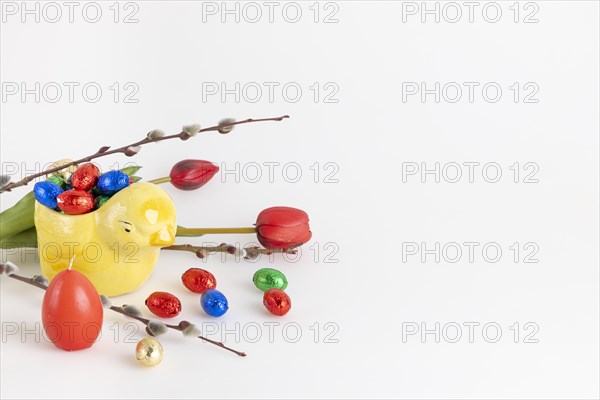 Colourful chocolate eggs in a yellow ceramic chick next to palm catkins, tulips and candle, white