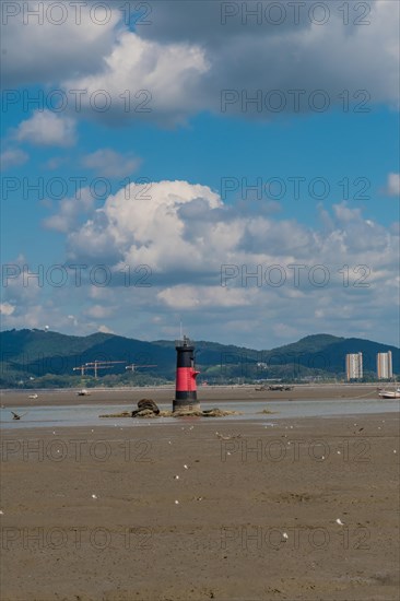 Red and black lighthouse on tidal flats during low tide with mountains and cloudy blue sky in