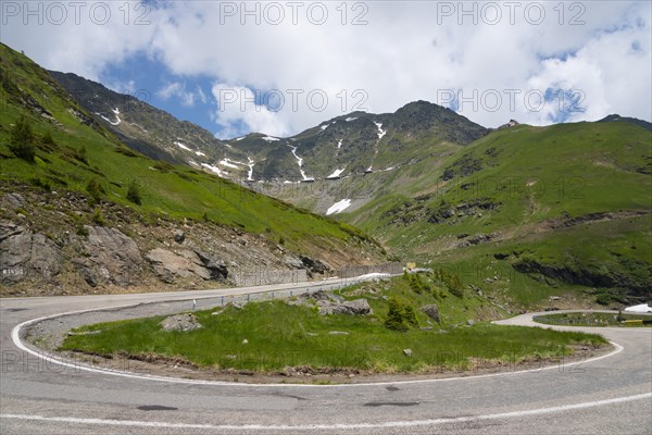 Fresh green hills and winding mountain road, partly covered by snow under a partly cloudy sky,