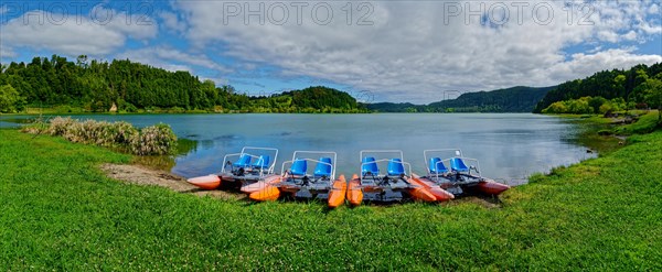 Tranquil Furnas Lake with pedal boats on the shore, surrounded by green nature and a cloudy sky,