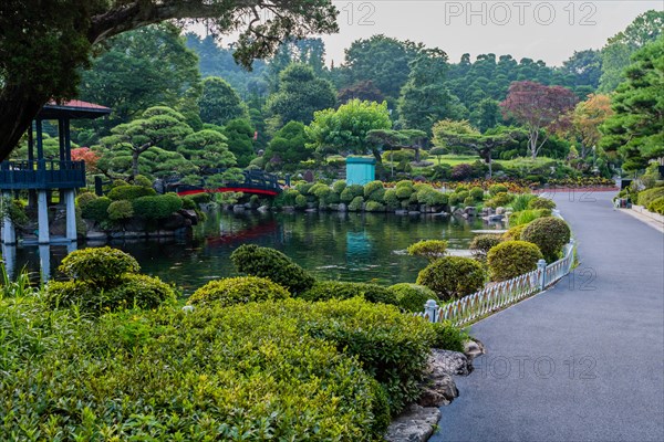 Paved walkway next to koi pond with a footbridge leading to gazebo in manicured public park in