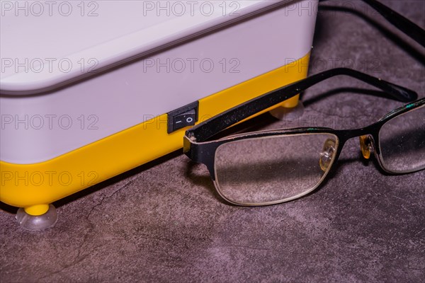Closeup of pair of wire framed glasses in front of ultrasonic cleaner machine on black textured