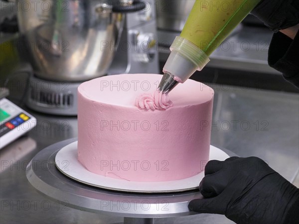 Close-up of a confectioner's piping bag adding pink frosting to decorate a cake with swirls,