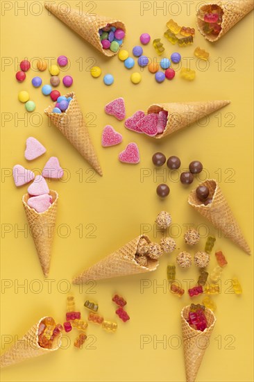 Colourful sweets, gummy bears and chocolate lentils fall out of ice cream cones on a yellow