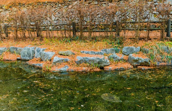 Boulders on shoreline of small frozen pond with wooden railing fence and stone wall in background