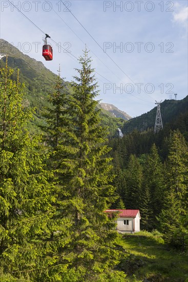 Small cable car gondola crosses a picturesque mountain landscape with fir trees and a small hut and