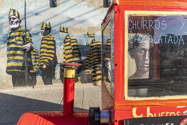 Red trolley for selling churros, behind a graffito with men in convict clothing, Ushuaia, Tierra
