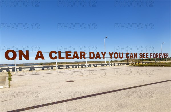 Large red rusty capital letters ON A CLEAR DAY YOU CAN SEE FOREVER blue sky background, Alqueva