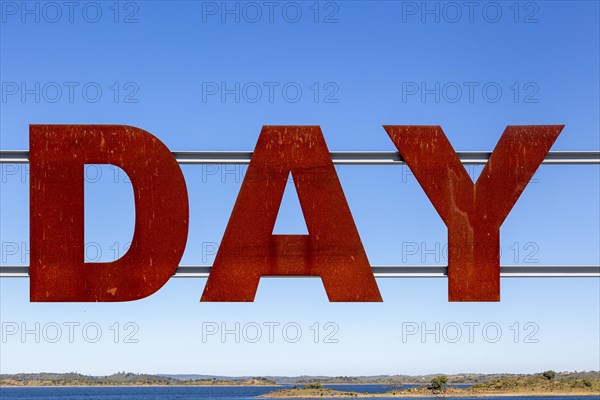 Large red rusty capital letters spell the word DAY against blue sky background, Alqueva dam, Moura,