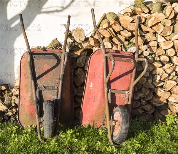 Two old red metal wheelbarrows leaning against pile wood logs in garden, Cherhill, Wiltshire,