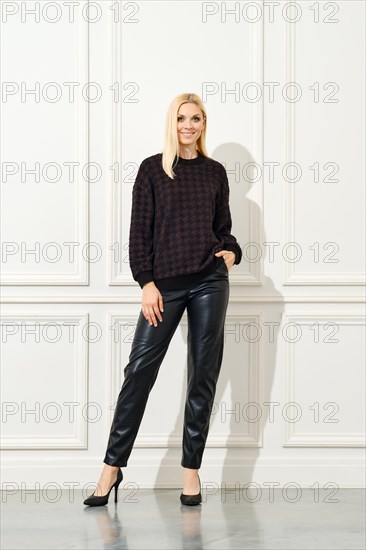 An elegant woman poses confidently against a white, panelled wall. She is dressed in a dark,