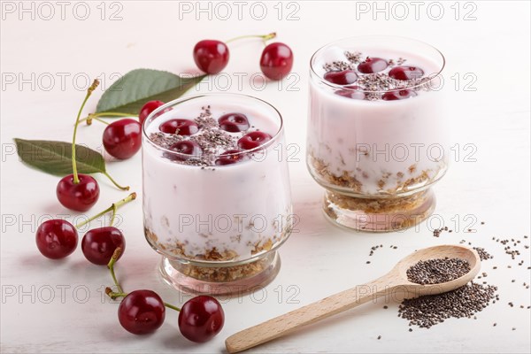 Yoghurt with cherries, chia seeds and granola in glass with wooden spoon on white wooden background