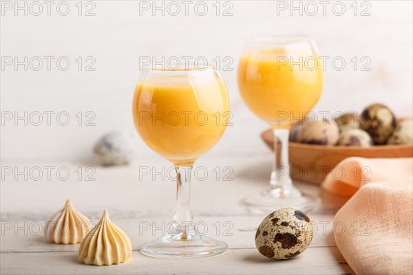 Sweet egg liqueur in glass with quail eggs and meringues on white wooden background. Side view,