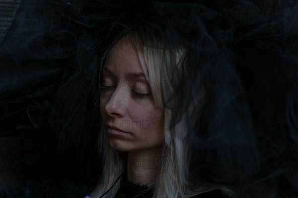 A young caucasian blonde woman's face is partially obscured by a dark veil, her eyes closed in a