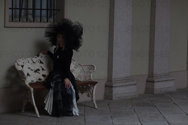 Thoughtful woman in black gothic attire sitting on a bench by a historical building, wear lace veil