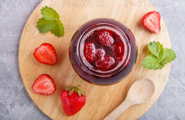 Strawberry jam in a glass jar with berries and leaves on gray concrete background. Homemade, top