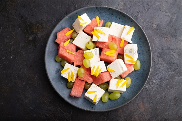 Vegetarian salad with watermelon, feta cheese, and grapes on blue ceramic plate on black concrete