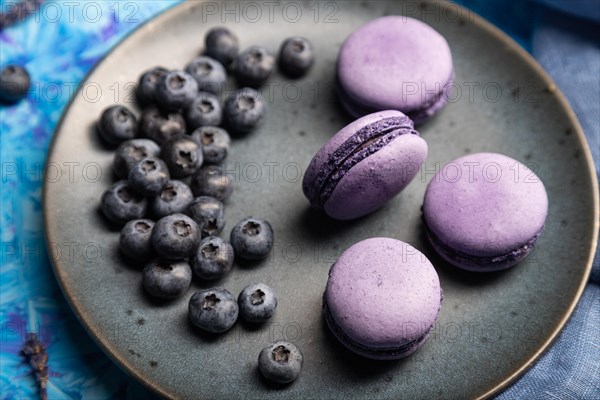 Purple macarons or macaroons cakes with blueberries on ceramic plate on a blue concrete background