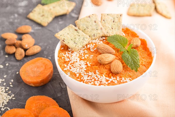 Carrot cream soup with sesame seeds, almonds and snacks in white bowl on a black concrete