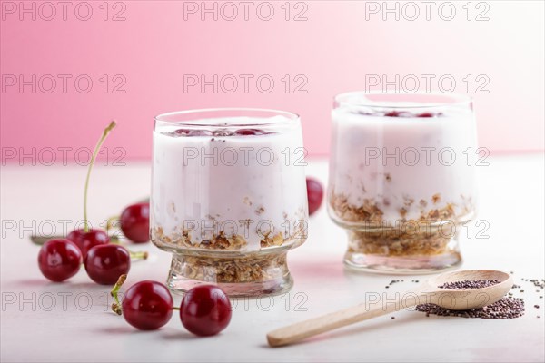 Yoghurt with cherries, chia seeds and granola in glass with wooden spoon on pink and white