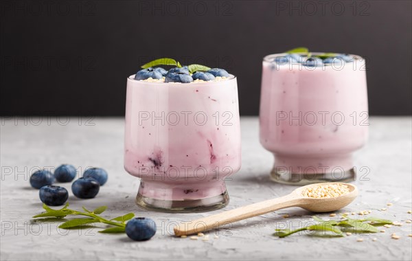 Yoghurt with blueberry and sesame in a glass and wooden spoon on gray and black background. side