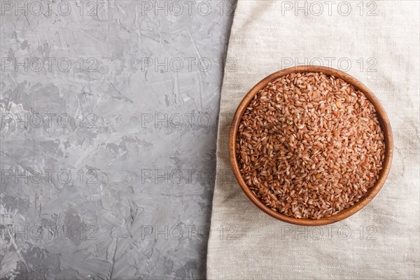 Wooden bowl with unpolished brown rice on a gray concrete background and linen textile. Top view,