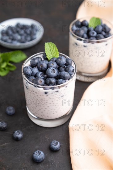 Yogurt with blueberry and chia in glass on black concrete background and orange linen textile. Side