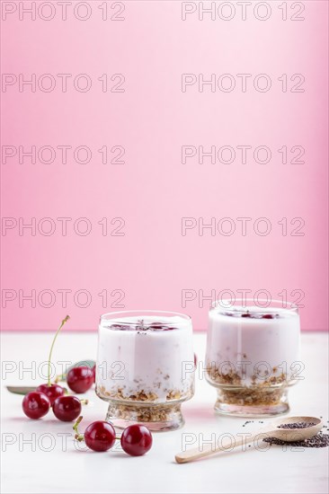 Yoghurt with cherries, chia seeds and granola in glass with wooden spoon on pink and white