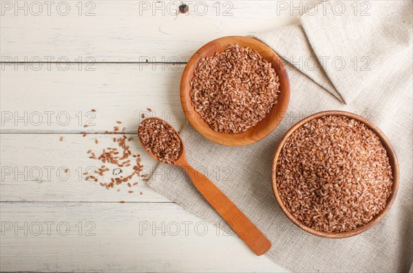 Two wooden bowls with unpolished brown rice and wooden spoon on a white wooden background and linen