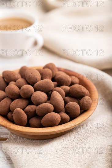 Almond in chocolate dragees in wooden plate and a cup of coffee on white wooden background and