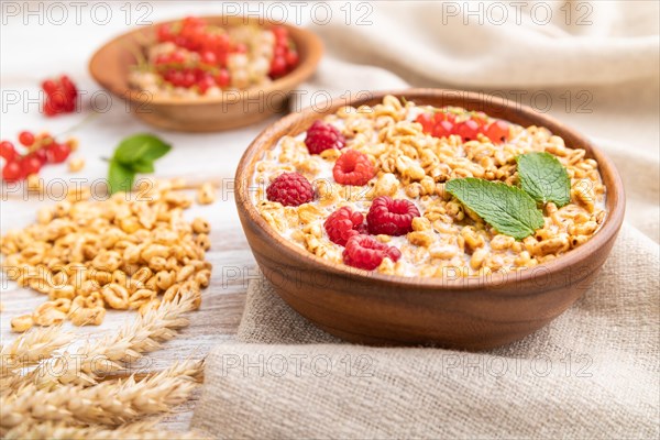 Wheat flakes porridge with milk, raspberry and currant in wooden bowl on white wooden background