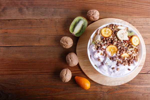 Vegetarian salad of bananas, apples, pears, kumquats, kiwi with granola and yogurt on brown woodem