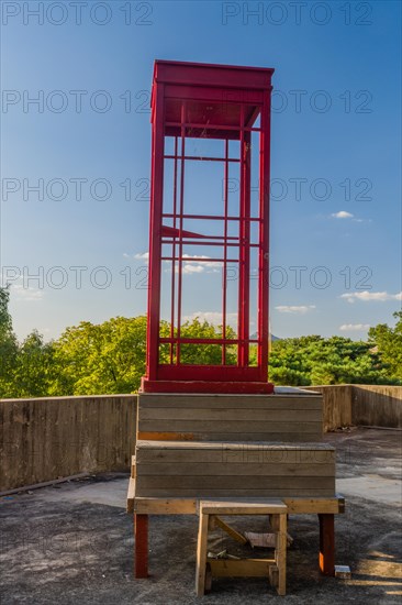 Red wooden telephone booth minus phone and without glass on roof of building on sunny day with blue