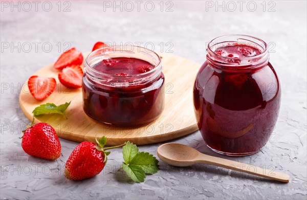 Strawberry jam in a glass jar with berries and leaves on gray concrete background. Homemade, close