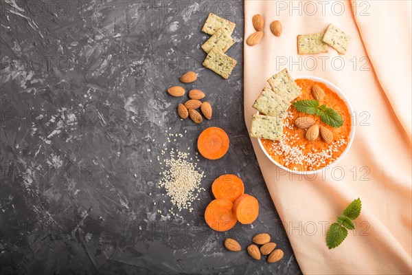 Carrot cream soup with sesame seeds, almonds and snacks in white bowl on a black concrete