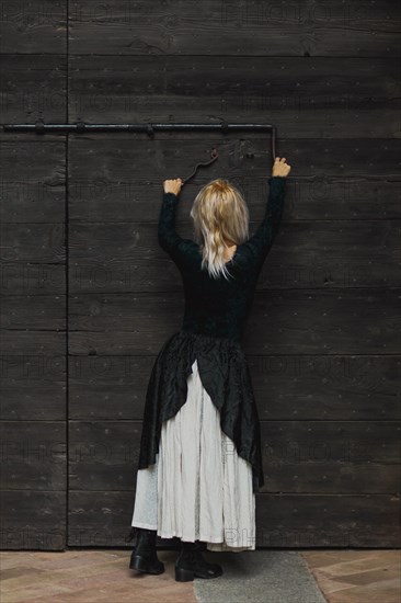 Full body shot of a young blonde caucasian woman reaching an antique lock of a wooden door above