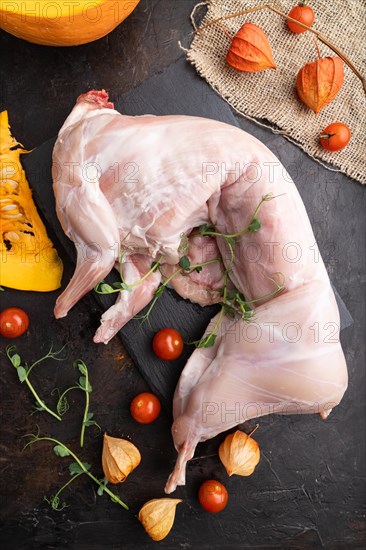 Whole raw rabbit with pumpkin, tomatoes, pea sprouts on a black concrete background and linen