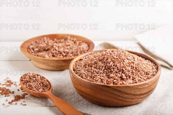 Two wooden bowls with unpolished brown rice and wooden spoon on a white wooden background and linen