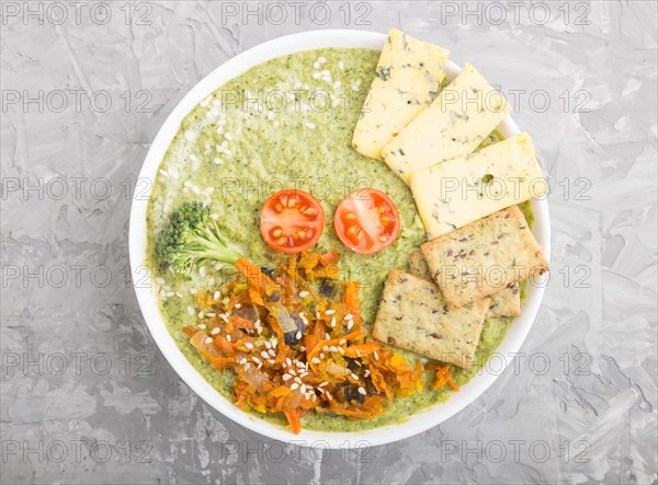 Green broccoli cream soup with crackers and cheese in white bowl on a gray concrete background. top