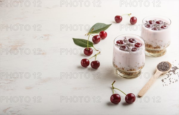 Yoghurt with cherries, chia seeds and granola in glass with wooden spoon on white wooden background