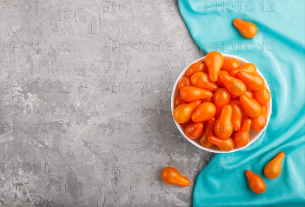 Fresh orange grape tomatoes in white ceramic bowl on gray concrete background with blue textile.