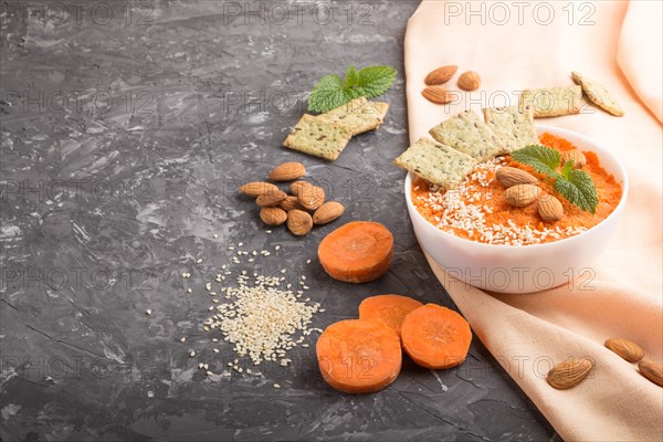Carrot cream soup with sesame seeds, almonds and snacks in white bowl on a black concrete