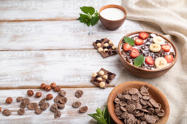 Chocolate cornflakes with milk and strawberry in wooden bowl on white wooden background and linen